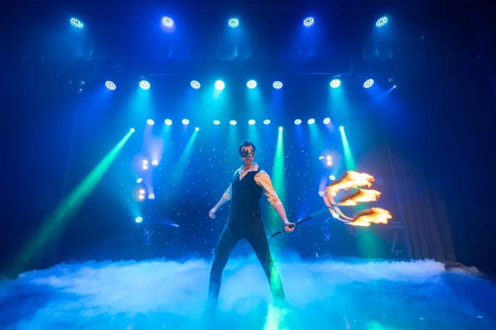 Man performing with flaming torches on stage with blue and green lights.