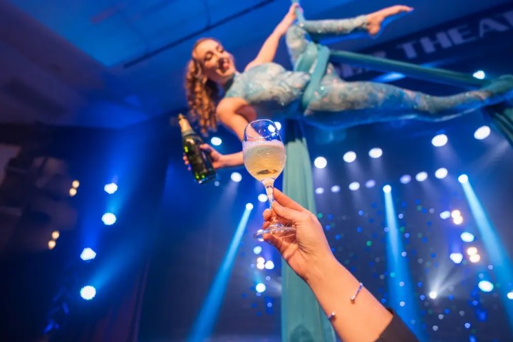 Aerialist in blue costume pours champagne into a glass on stage with bright lights.