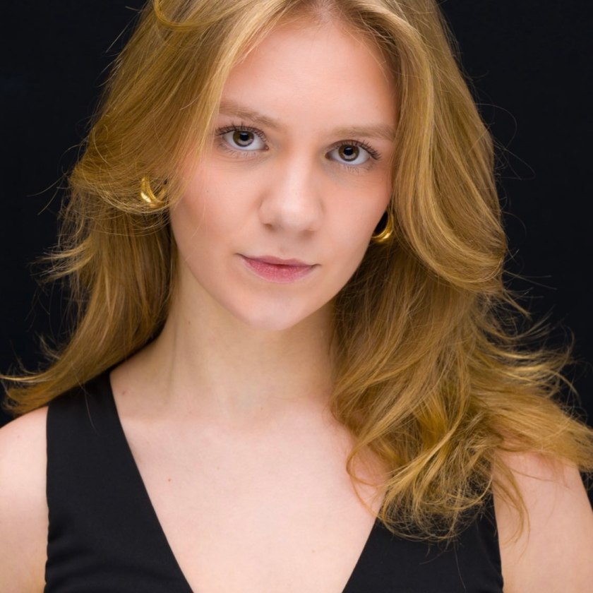 Young woman with wavy blonde hair and gold earrings wearing a black top.