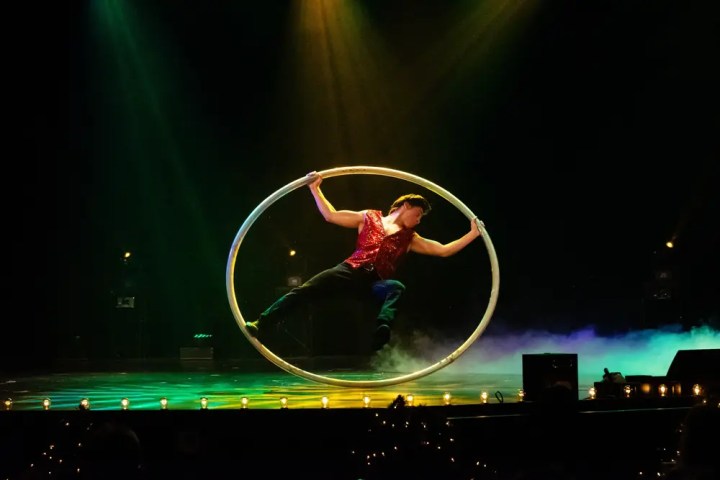 Performer in a red shirt balancing inside a large hoop on a dimly lit stage.