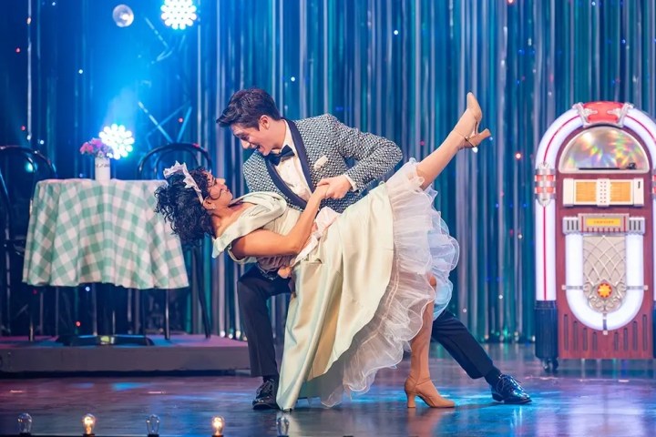 Couple in 1950s attire dancing with jukebox and table in background.