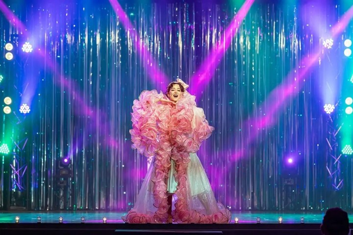 Performer in a pink, ruffled dress on stage with colorful lights and tinsel backdrop.