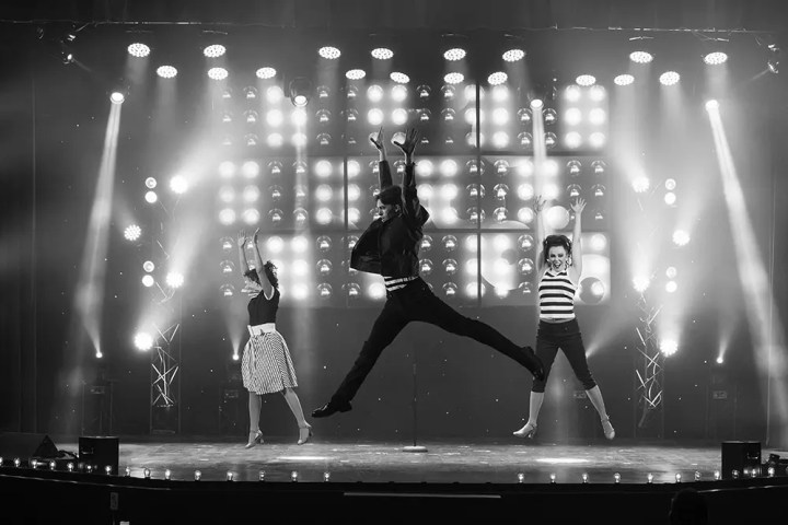Three dancers energetically perform on a lit stage with spotlights in a black-and-white photo.