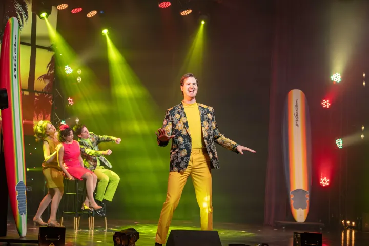 Performer in yellow suit sings on stage with people seated, colorful lights, and surfboards.
