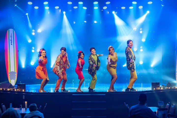 Six performers in colorful outfits dance on a stage with bright blue lights.