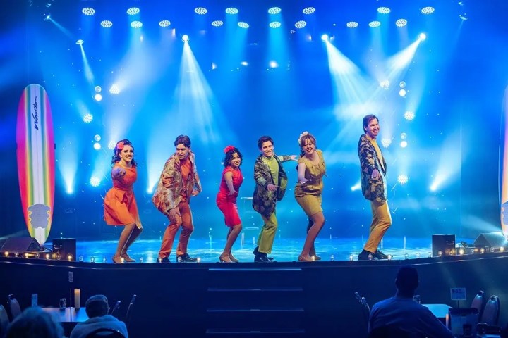 Six performers in colorful outfits dance on stage with bright lights and surfboards.