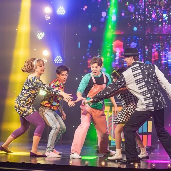 Performers in colorful outfits dance energetically on stage with vibrant lighting.