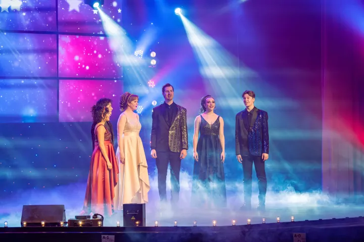 Five people in formal wear perform on a stage with colorful lights and fog.