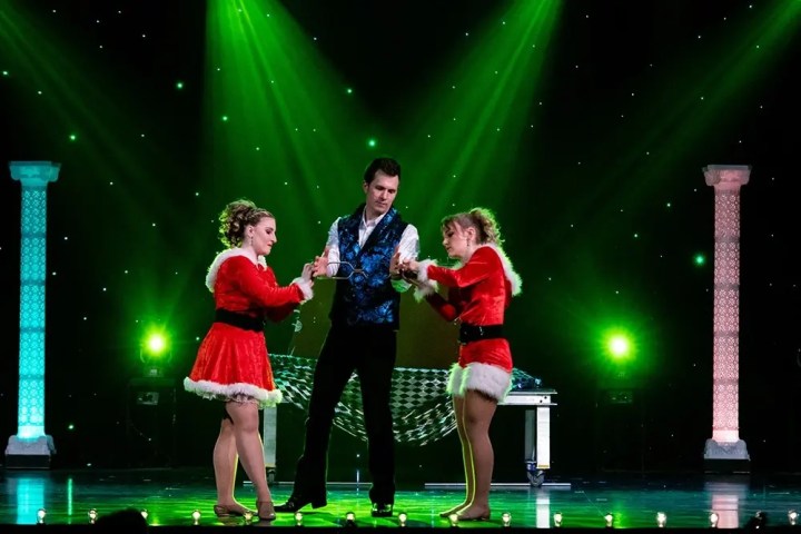Magician and two assistants in red costumes perform under green lights on stage.