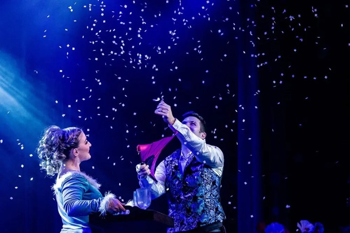 Two magicians performing with confetti and red cloth on a dimly lit stage.