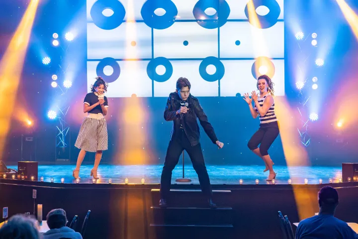 Three performers on stage, one singing with a mic, colorful lights in the background.