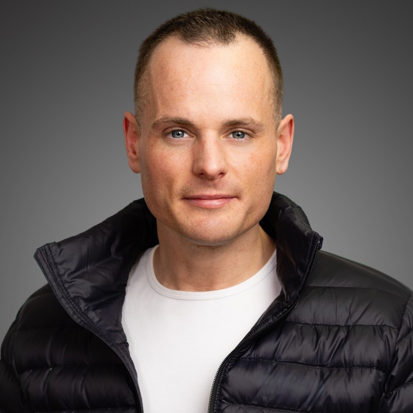Man in black jacket and white shirt against dark background.