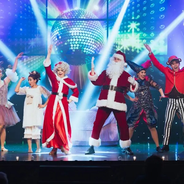 Performers in colorful costumes, including Santa, dance on stage with a disco ball and festive lights.