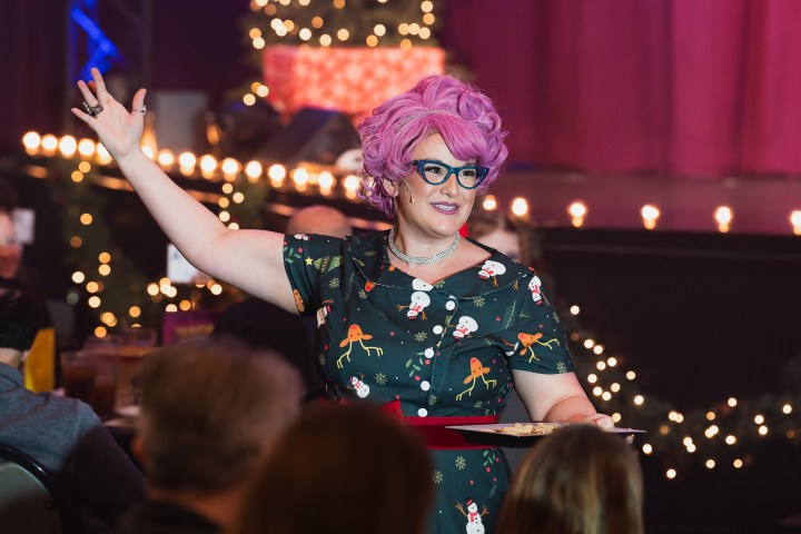 Person with pink hair and festive dress gestures on stage with audience in background.