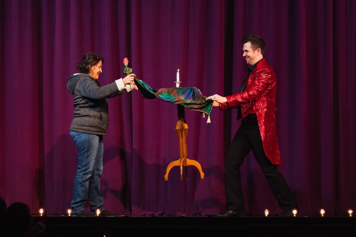 A magician in a red jacket performs a levitation trick with a woman on a stage.