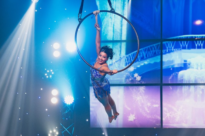Aerial performer in blue outfit hanging from a hoop with stage lights and snowy backdrop.
