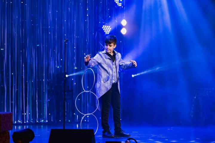 Young magician on stage performing with rings, blue lighting and shimmering backdrop.