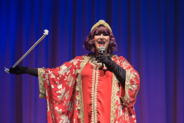 Performer in red costume with microphone and wand on stage.
