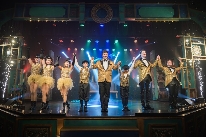 Performers in gold costumes take a bow on a theater stage with colorful lights and sparkles.