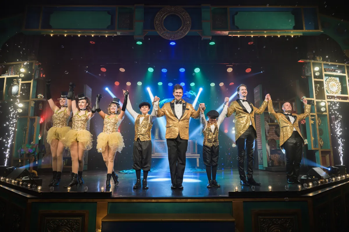 Performers in gold costumes take a bow on a theater stage with colorful lights and sparkles.