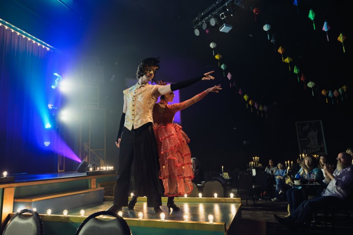 Stage performance with two dancers in costumes, audience watches from tables with hanging lanterns overhead.