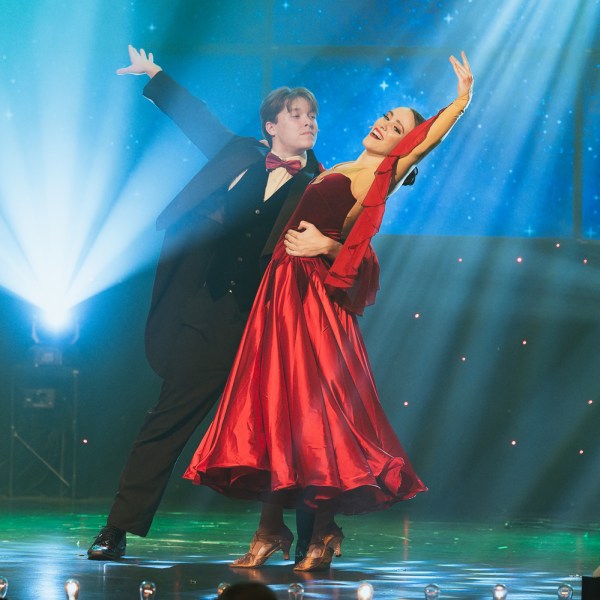 Elegant couple dancing in formal attire on a stage with blue lighting and starry backdrop.