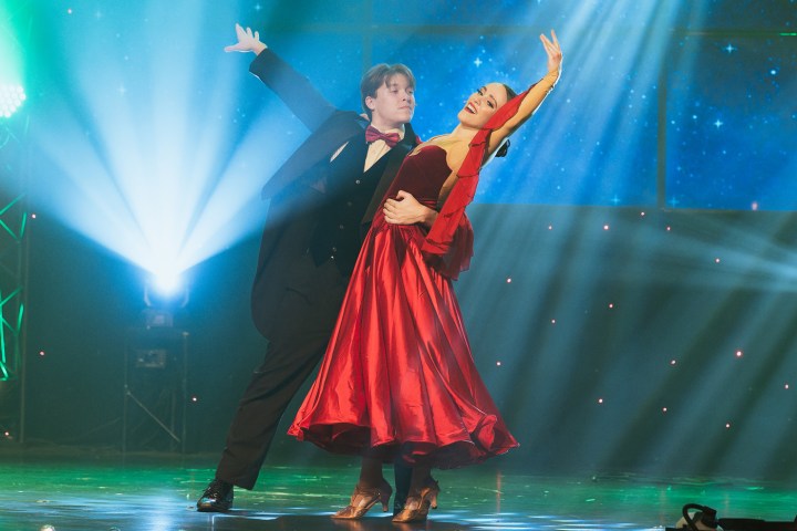 Elegant couple dancing in formal attire on a stage with blue lighting and starry backdrop.