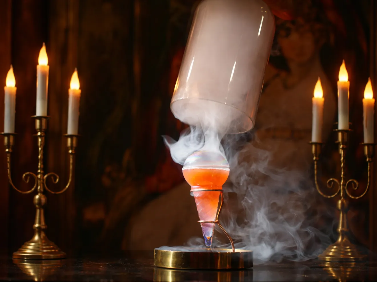 Two candelabras and a smoking cocktail glass under a glass dome on a table.