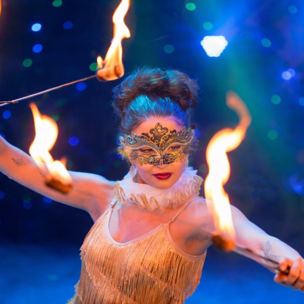 Performer in gold costume and mask twirling fire sticks on a colorful stage.
