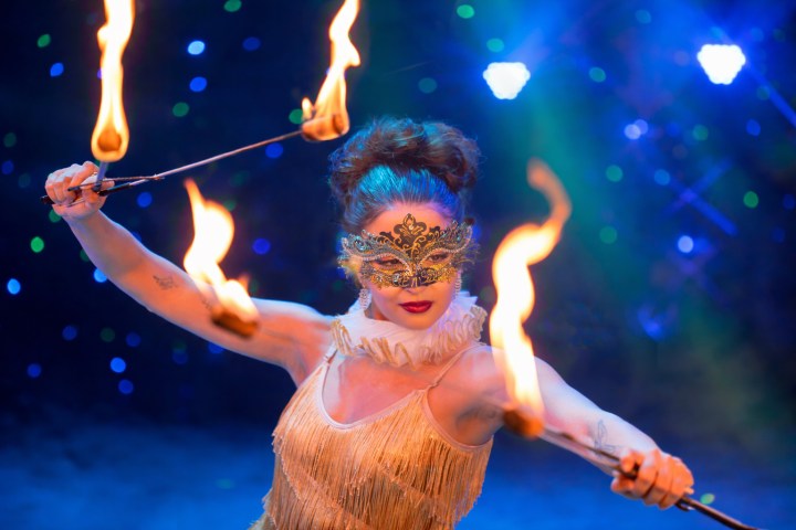 Performer in gold costume and mask twirling fire sticks on a colorful stage.