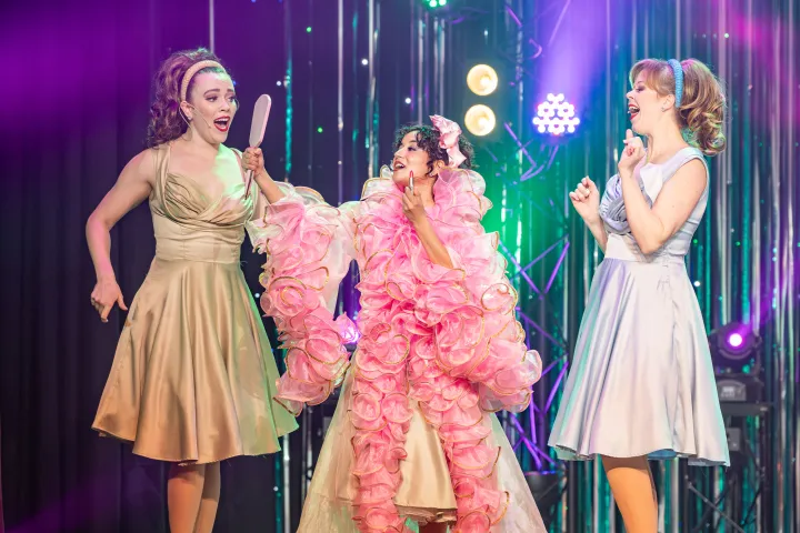 Three performers in colorful dresses sing on stage with bright lights.