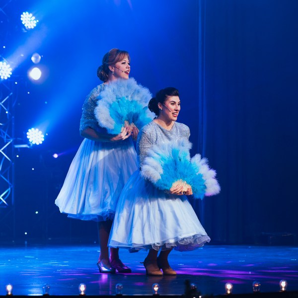 Two performers in blue dresses dancing with blue fans on a stage with colorful lights.