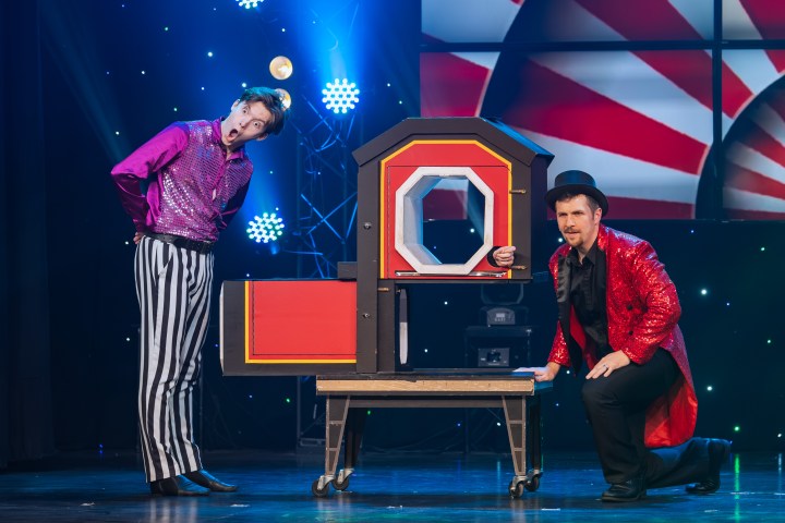 Two magicians performing an illusion with a box and a person inside, on stage with bright lights.