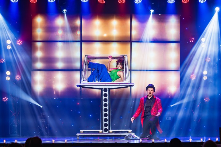 Magician on stage with assistant in a box, colorful lights illuminating the performance.