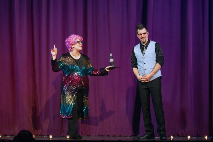 Two performers on stage, one in a colorful sequin coat with pink hair, holding a bottle; the other in a vest and tie.
