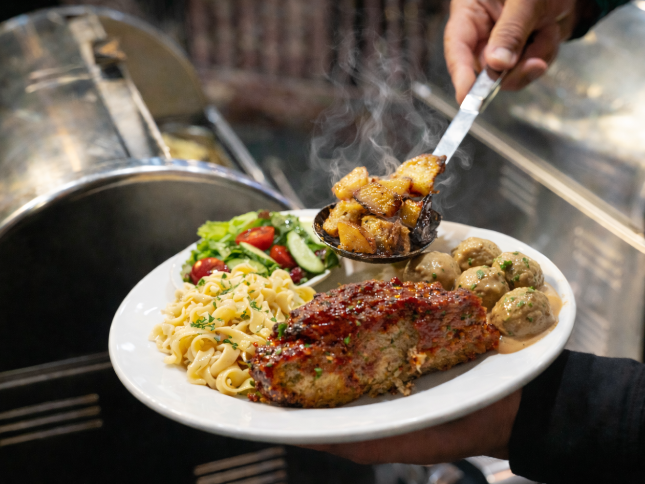 Plate of food with roast potatoes, salad, pasta, meatloaf, and meatballs, held by a person.