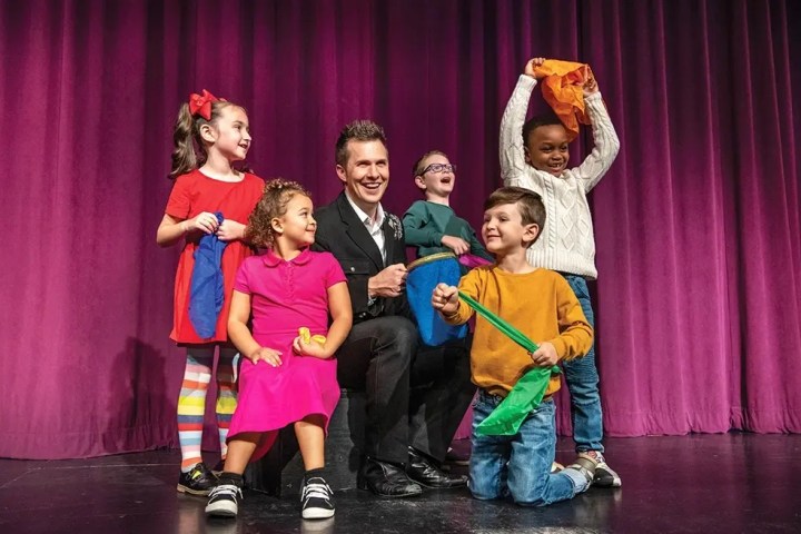 Magician with kids on stage, holding colorful scarves, against a purple curtain.