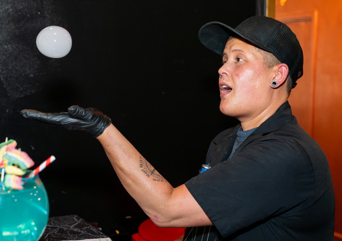 Person in black cap and gloves balances bubble on hand in a dim setting.