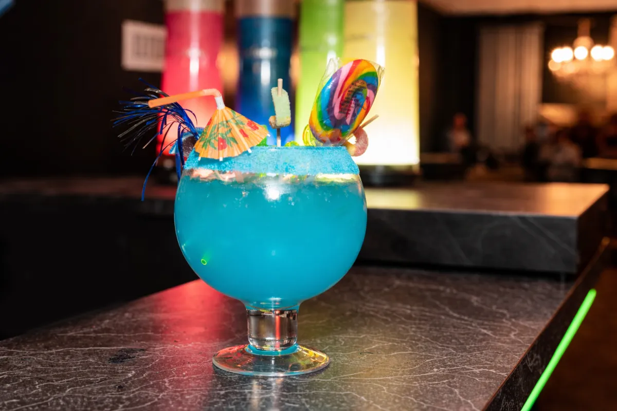 Bright blue cocktail in a globe glass with lollipop, straw, and mini umbrella.