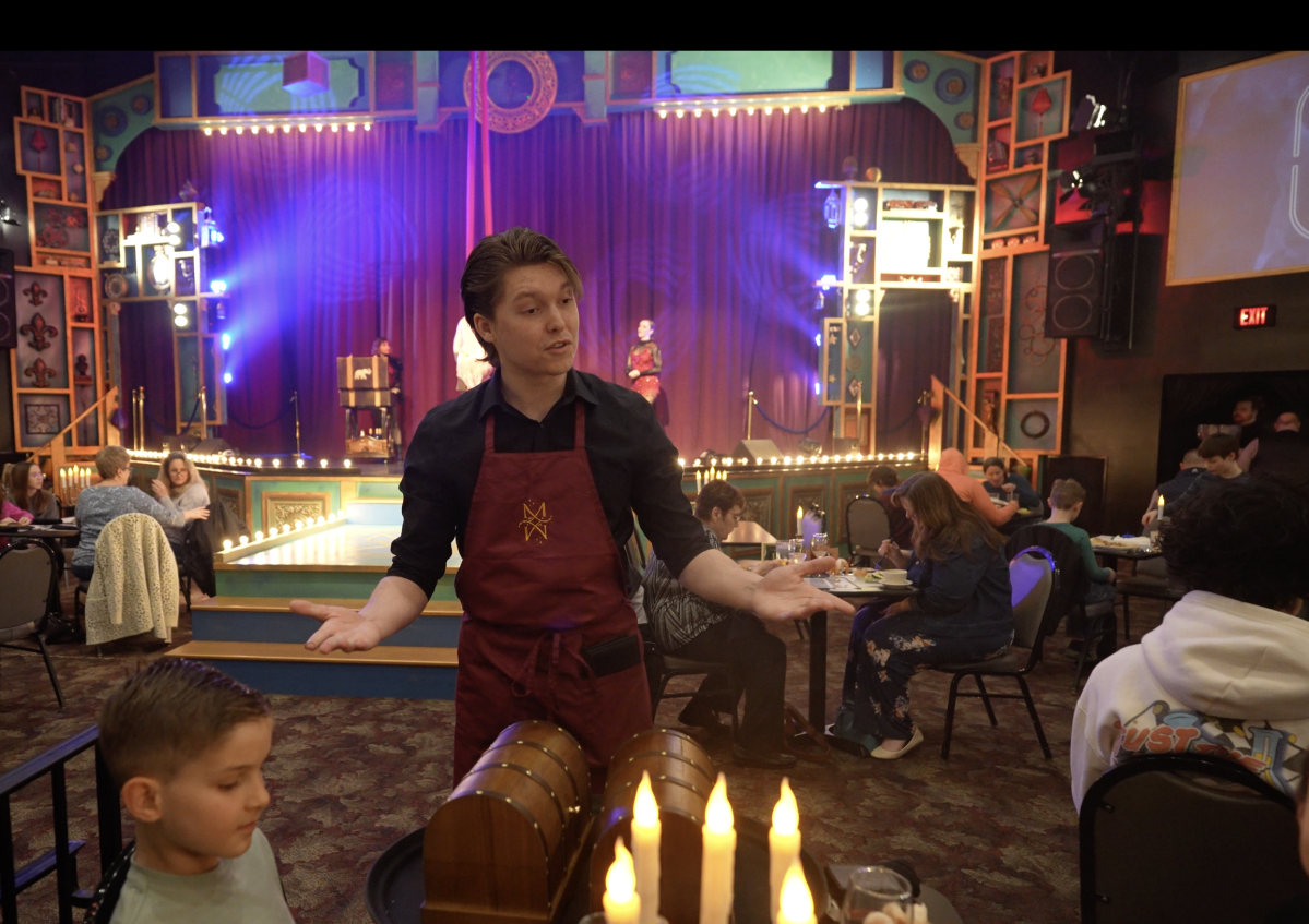 Performer in red apron gestures to audience in a colorful, lively theater setting.