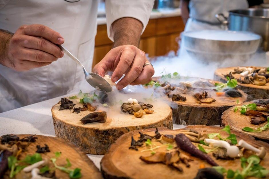 Chef plating food with steam on wooden boards in a kitchen.