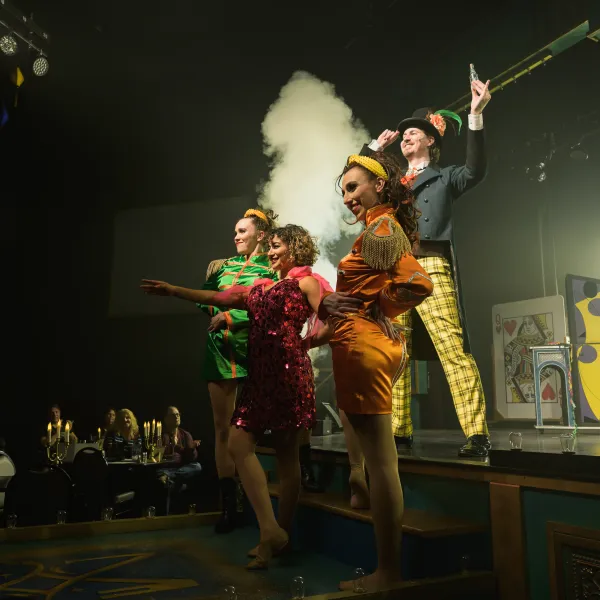 Performers in colorful costumes on stage with smoke and audience seated at tables.