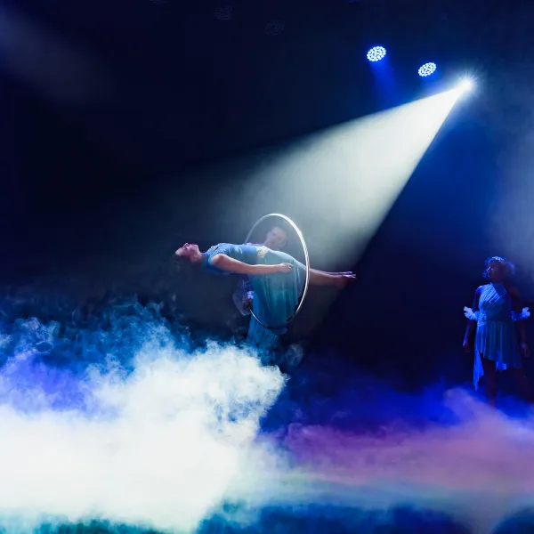 Performer hula-hooping in spotlight on stage with mist and spectators.