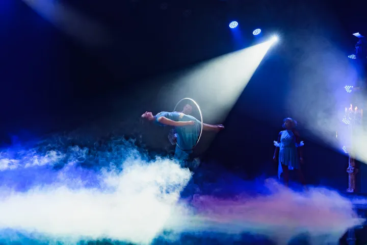 Performer hula-hooping in spotlight on stage with mist and spectators.