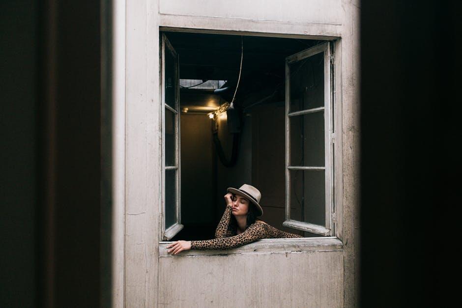 Woman in a hat leaning out of a window, with a pensive expression.