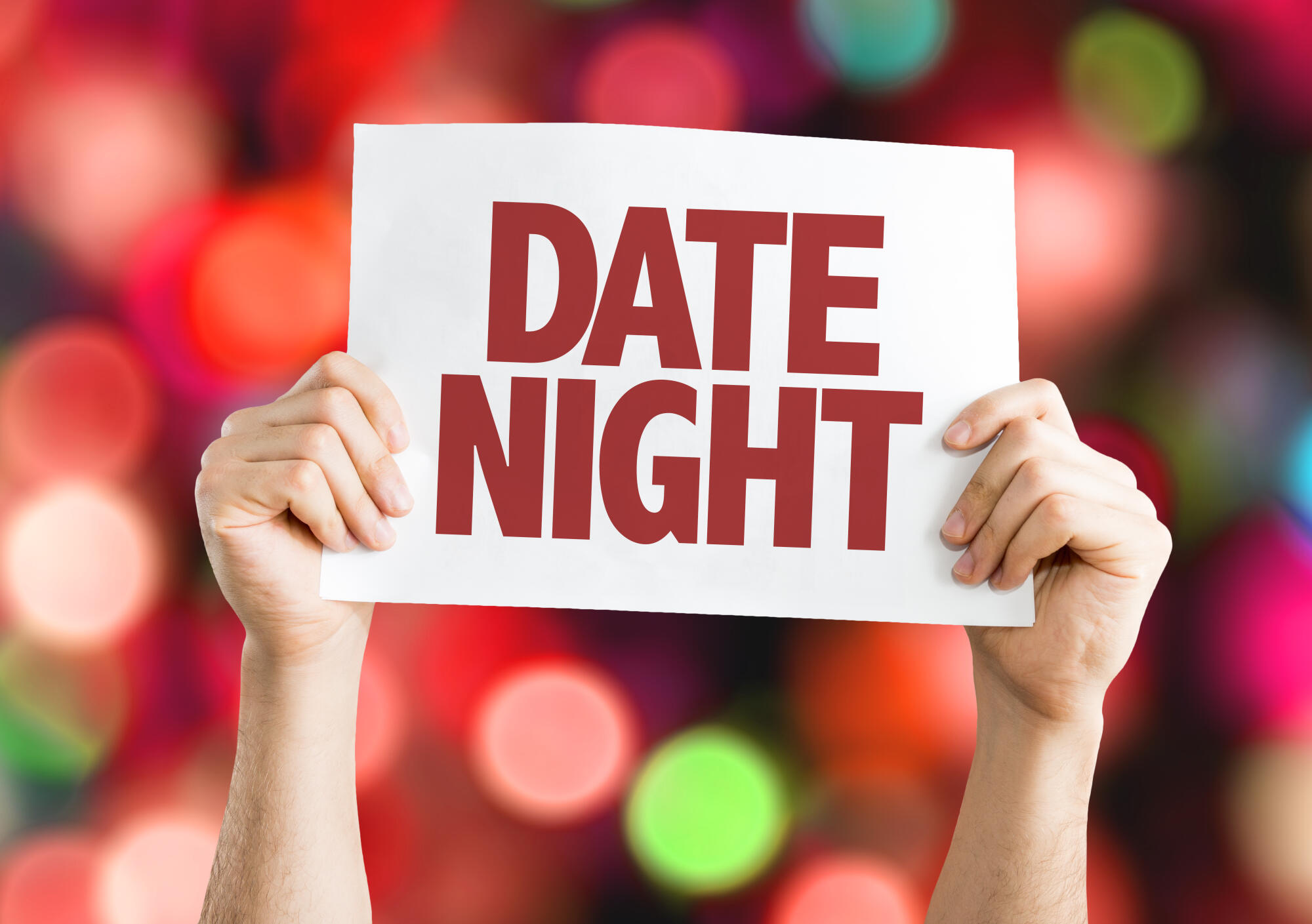 Hands holding a sign saying 'Date Night' with colorful blurred lights in background.