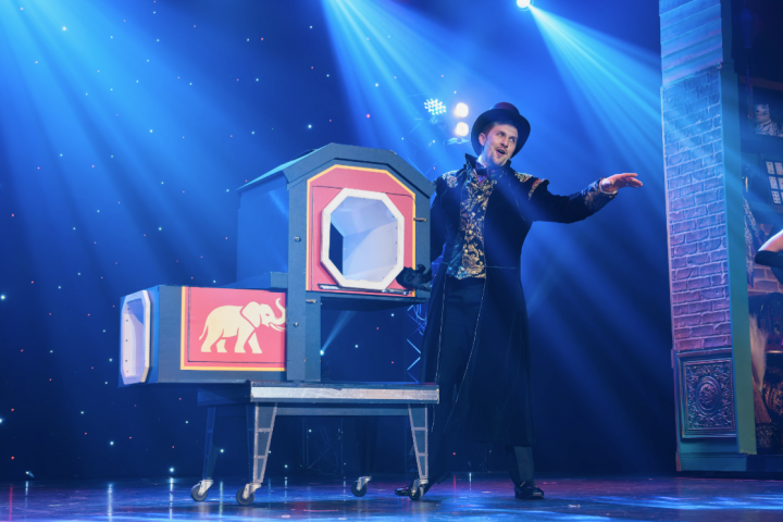 Magician performs with a box on stage, under blue lighting.