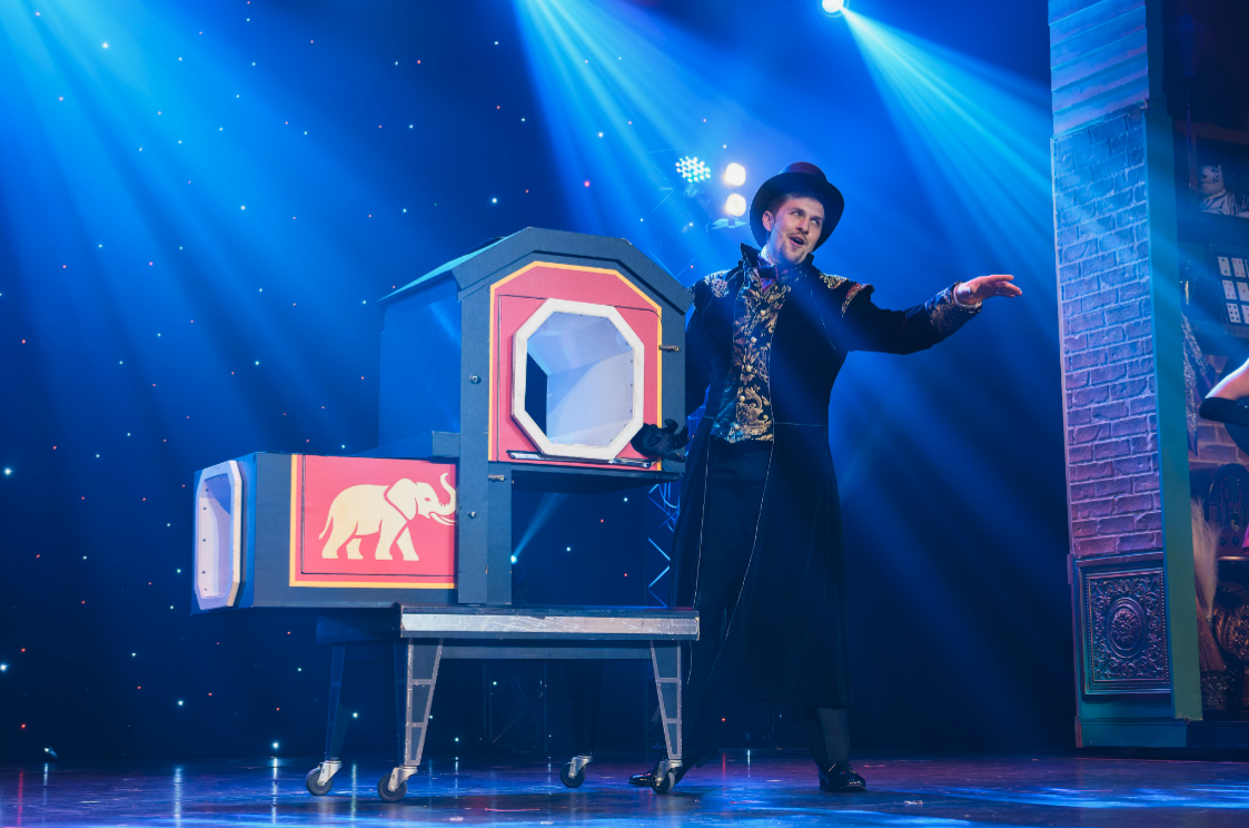 Magician performs with a box on stage, under blue lighting.