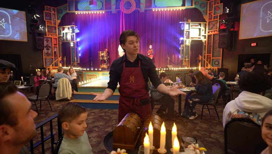Performer in red apron gestures on stage with audience seated around.