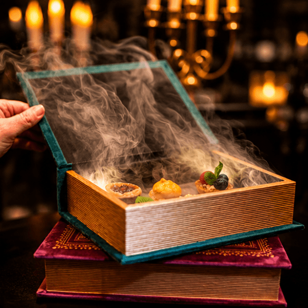 A hand opened a box styled as a book emitting smoke, revealing assorted gourmet dishes inside.